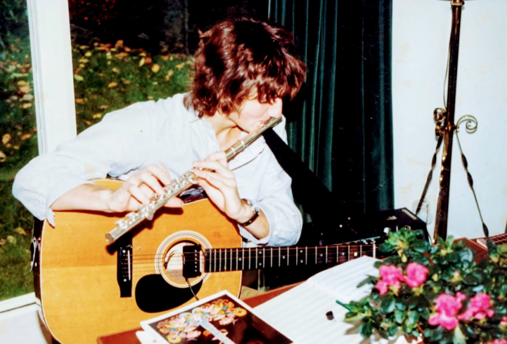A young man playing the flute with a guitar in his lap 
