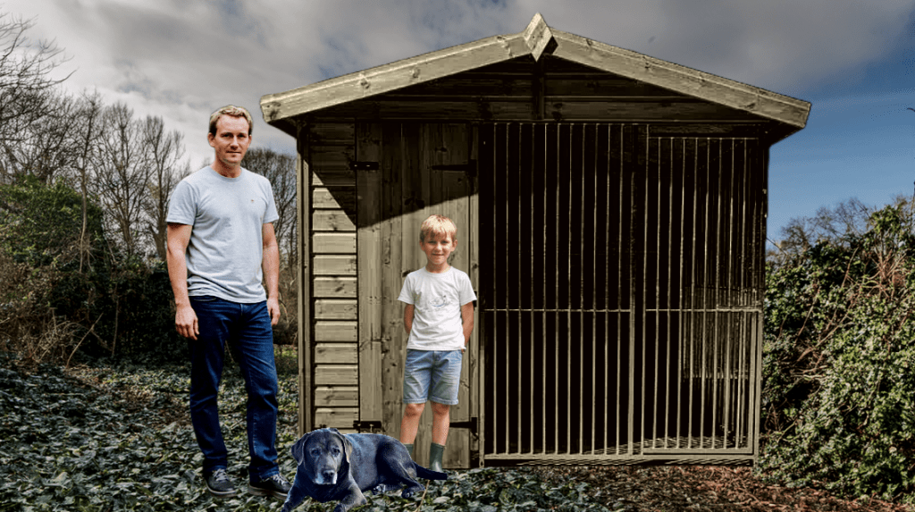 A man in his early thirties stands with a boy of 8 or 9 in front of a dog and a dog kennel in a patch of overgrown garden.