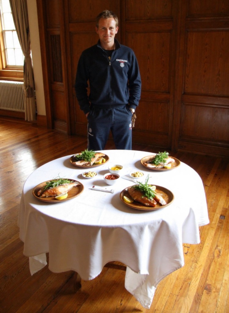 A person standing near a round table set with several dishes of poached salmon garnished with greens and accompanied by various sauces and condiments in a wooden room.