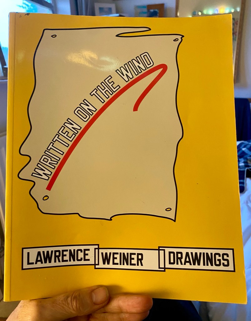 A hand holding a book titled 'Written on the Wind' by Lawrence Weiner, featuring a yellow cover with red and black text.