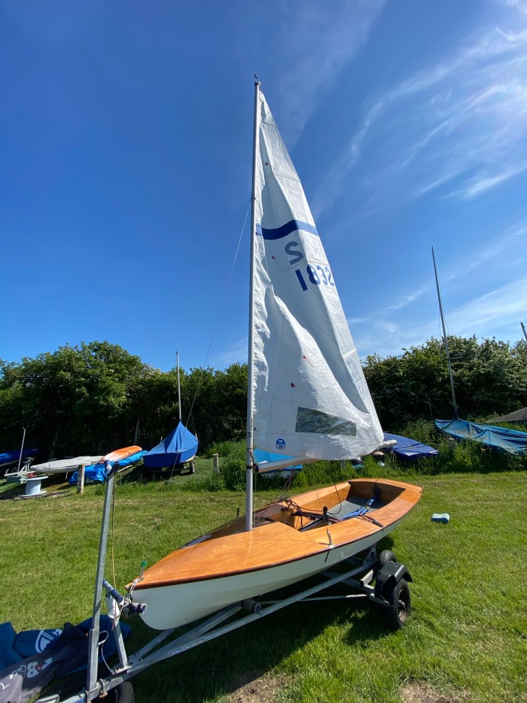 A wooden Streaker racing dinghy with a sail, parked on a grassy area surrounded by other boats.