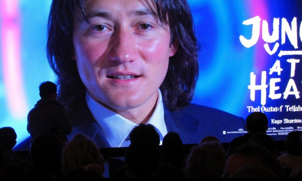 A large screen displays a close-up of a man with long hair, while an audience is silhouetted in the foreground. The text 'JUNGLE AT HEART' is prominently featured on the screen.