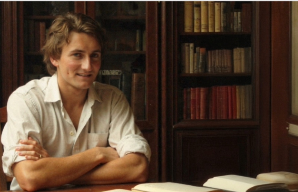 A young man with short, tousled hair sits at a table, smiling slightly. He is wearing a casual, light-colored shirt and is seated in front of a wooden bookshelf filled with books, suggesting a scholarly or creative atmosphere.