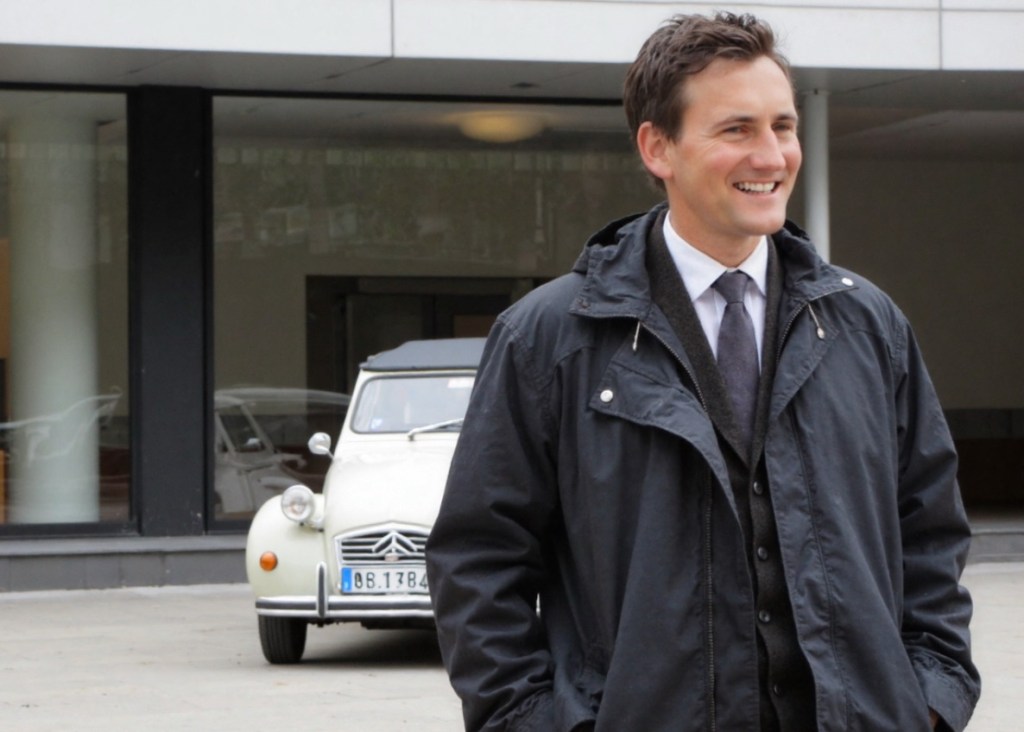A man in a black jacket stands smiling in front of a modern building, with a vintage Citroën 2CV car parked behind him.