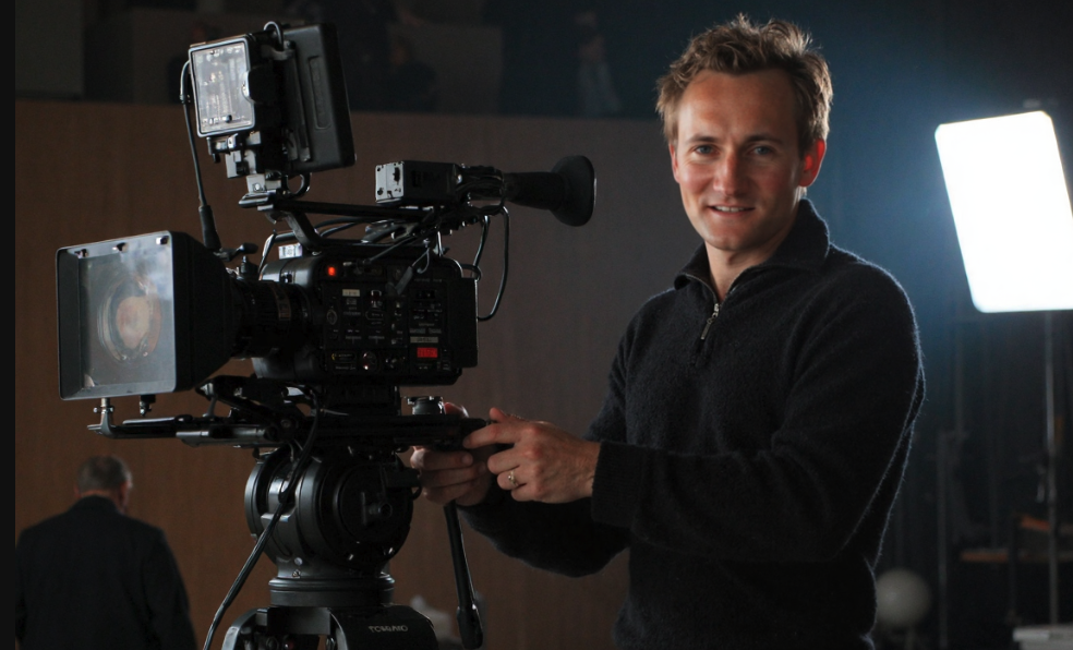A man smiling while holding a professional camera on a tripod, with studio lighting in the background.